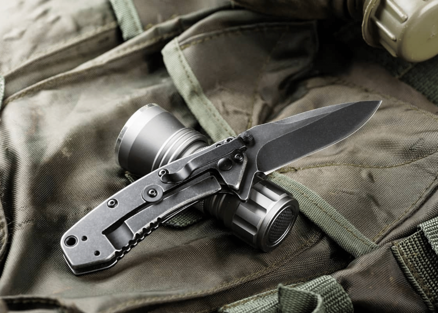 Black tactical knife, open, with a small flashlight on a dark backpack