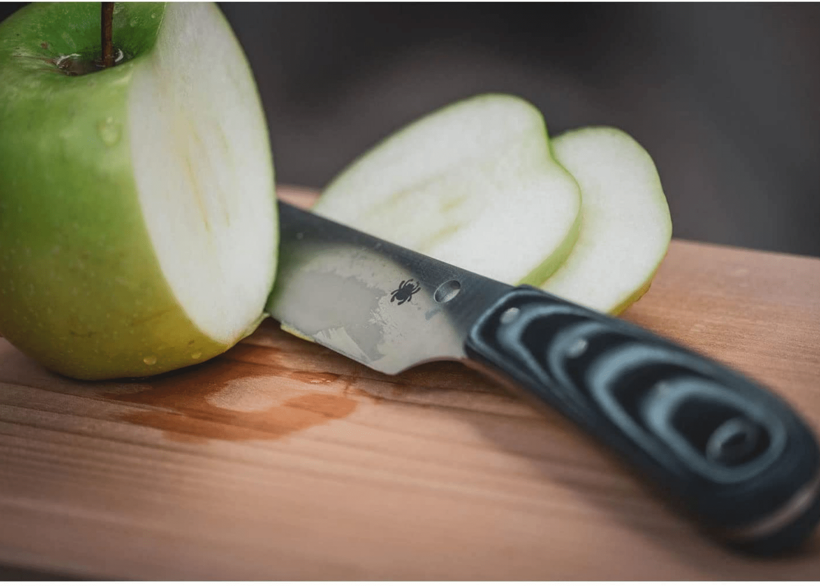 A Camp knife with a green apple cut in half on a red picnic table up close