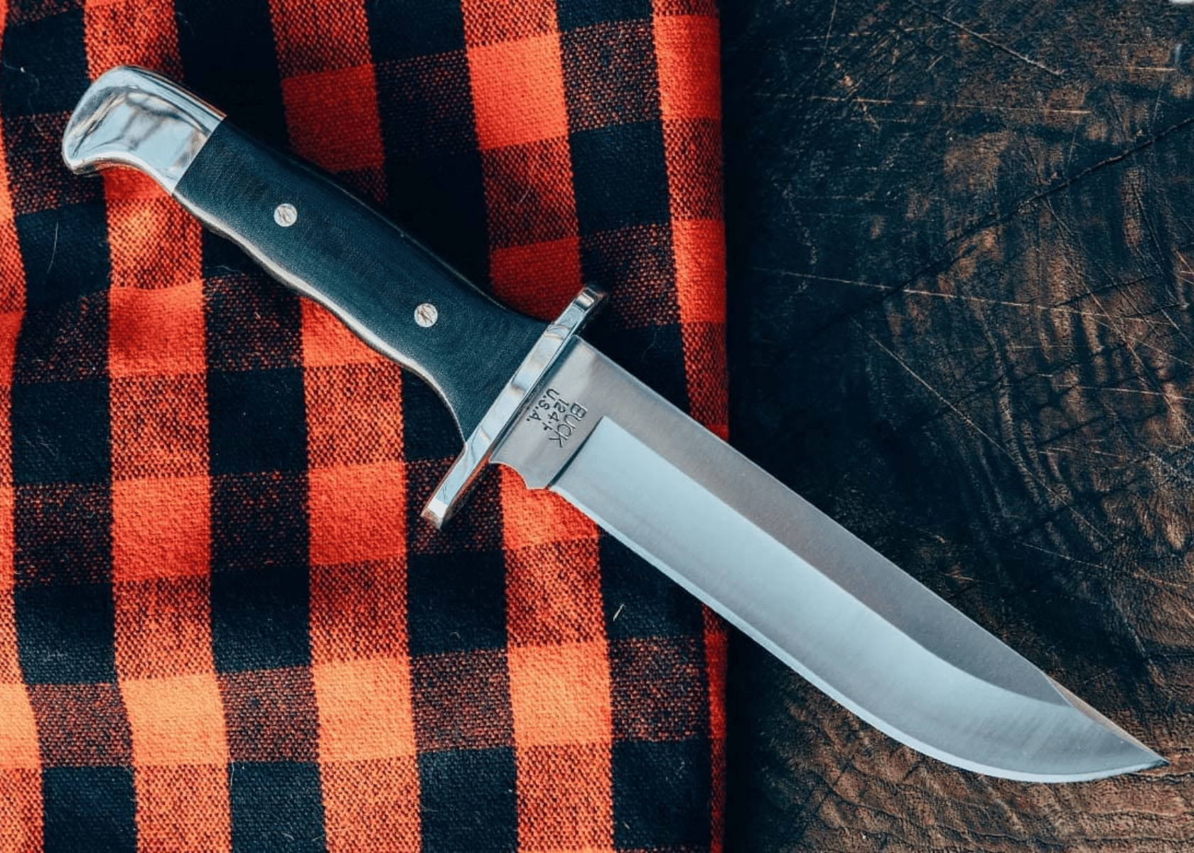 A Buck Knife laying on a red/black plaid cloth and wood