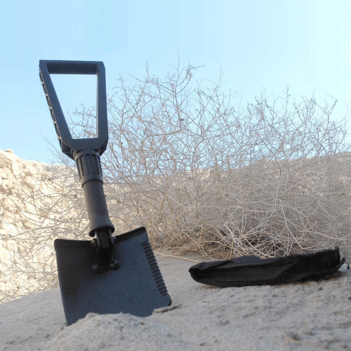 A survival shovel in the sand next to some dead brush