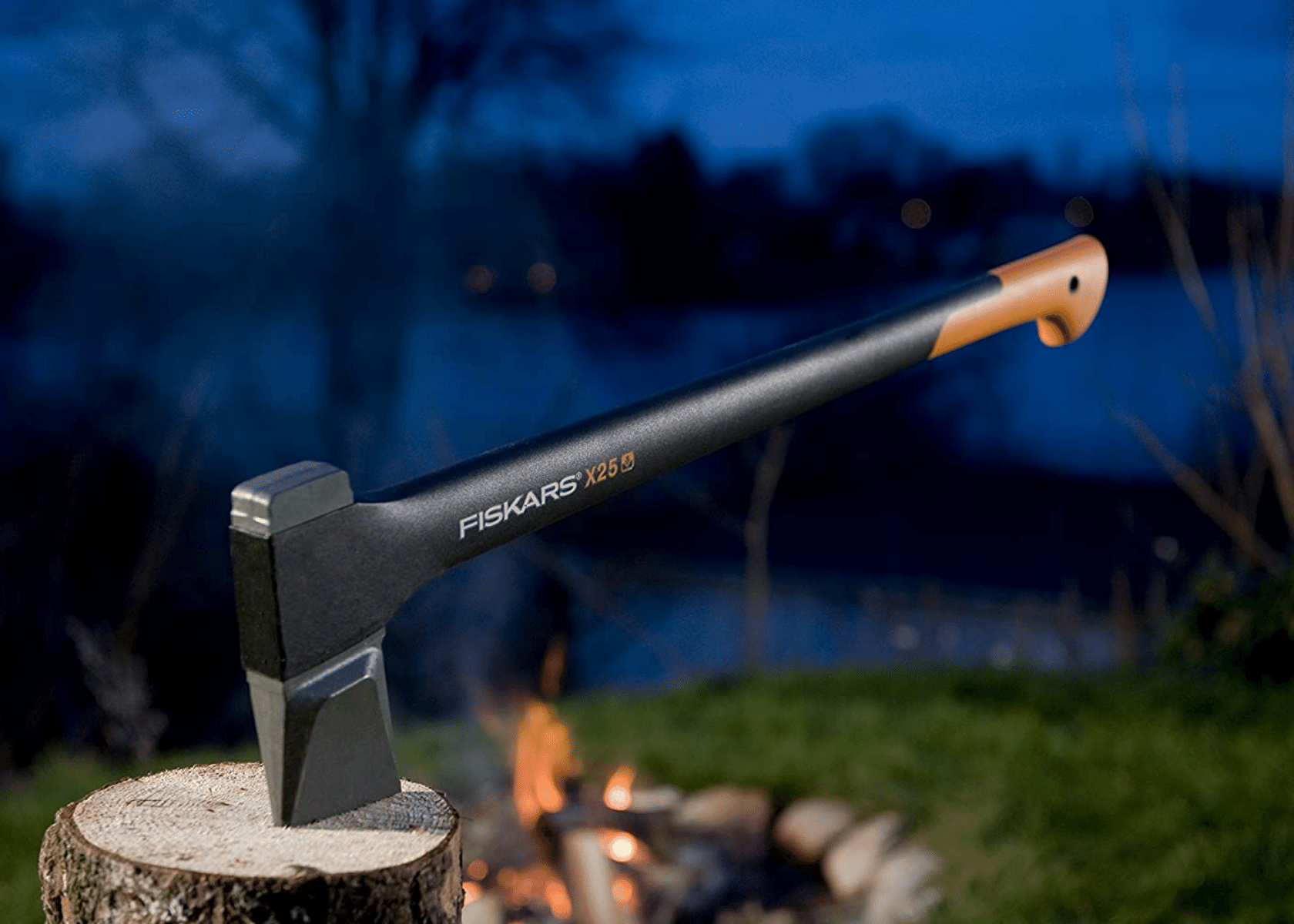 A wood-splitting axe stuck in the end of a piece of wood by a fire in a yard next to a lake