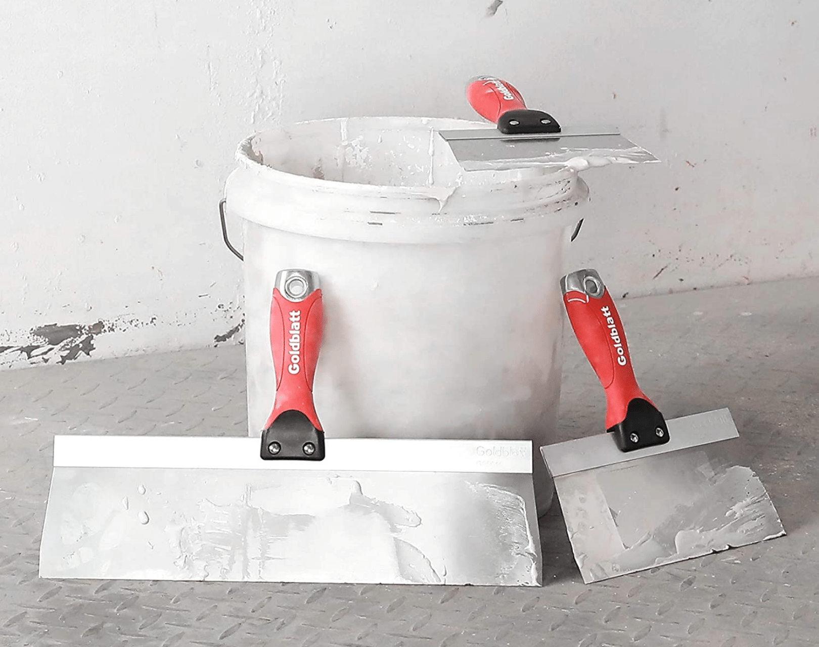 Two different size drywall knives leaning against a 5-gallon bucket of drywall mud