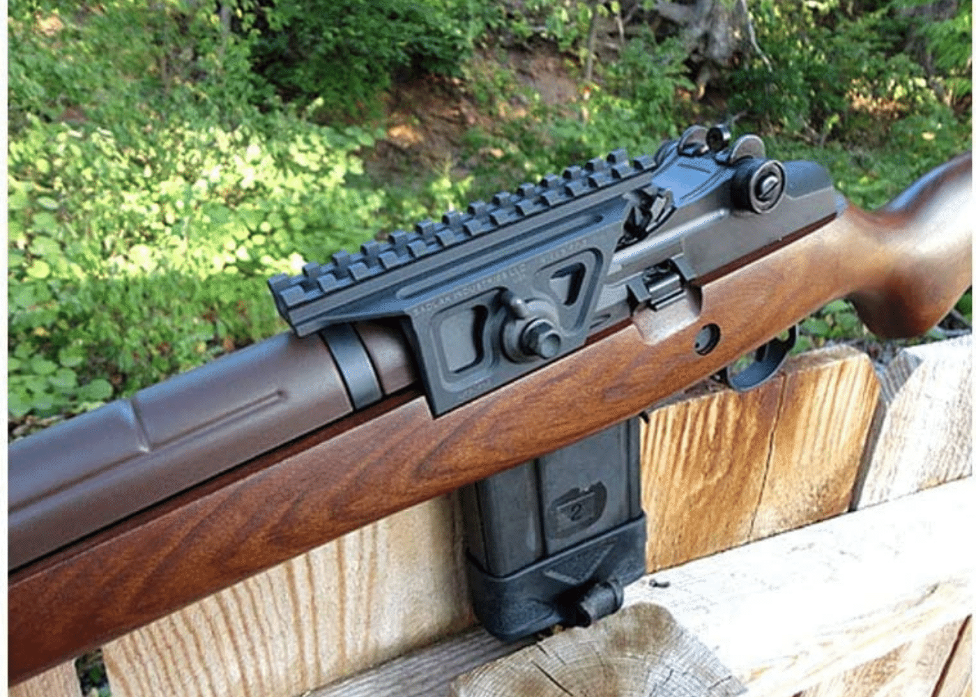 A M1A rifle with scope mount sitting on a wood fence with woods in the background