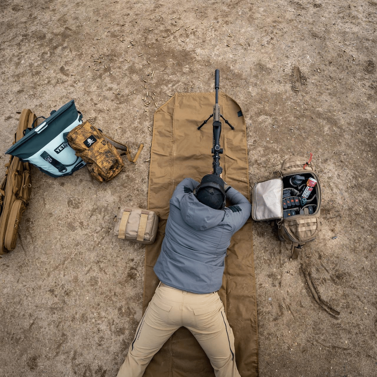 A man, prone on a coyote brown mat with his rifle in position and his range bag and gear around him