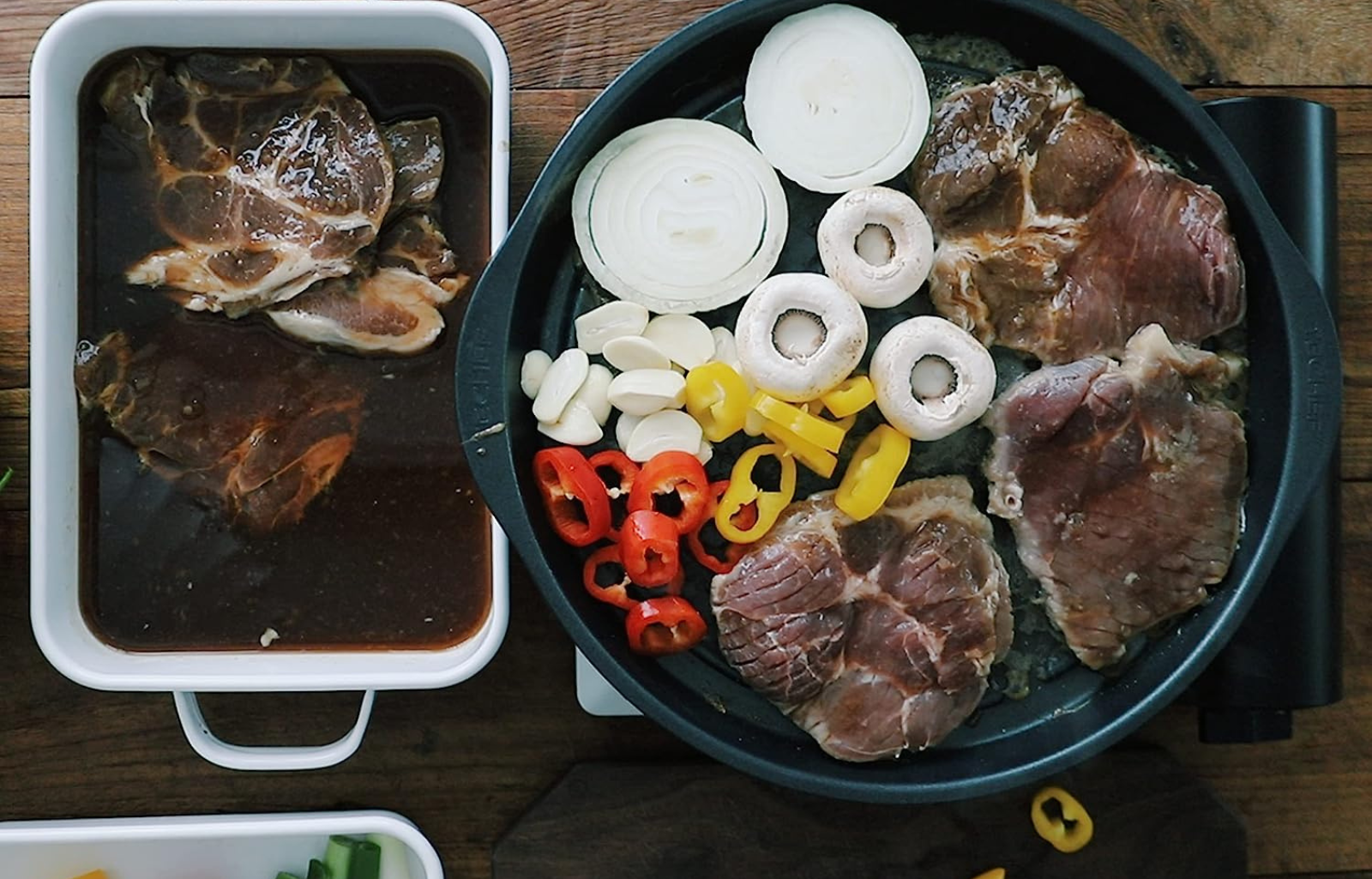 A Korean BBQ grill loaded with meat and vegetables with a dish beside it with more meat to be cooked