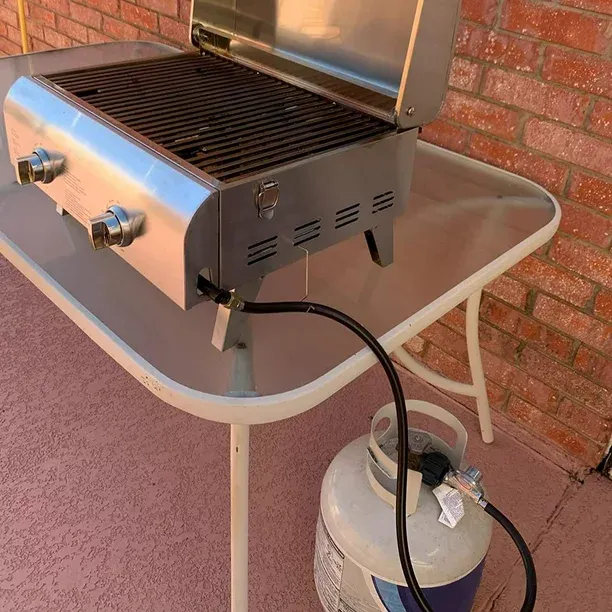 A tabletop grill with a gas grill regulator remotely attached to a propane tank under the glass table.