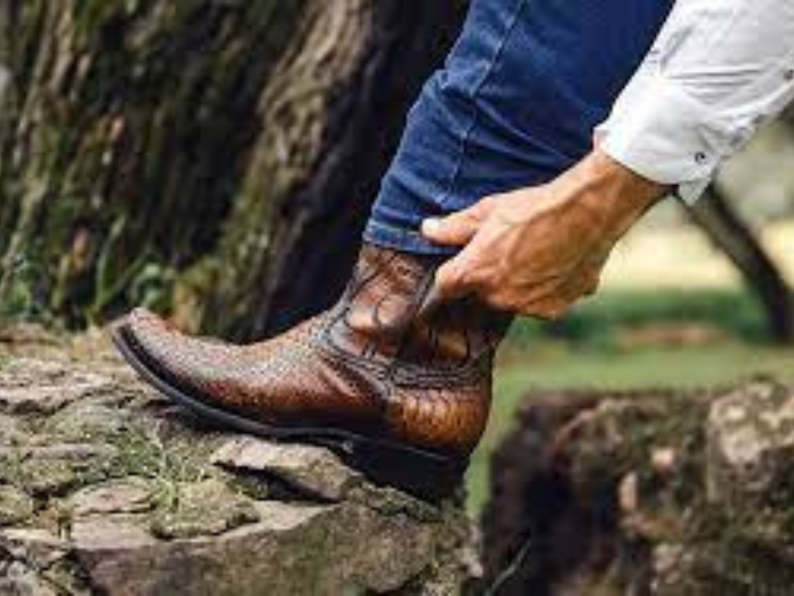 A man with his foot on a rock is pulling his jeans down over his boot.