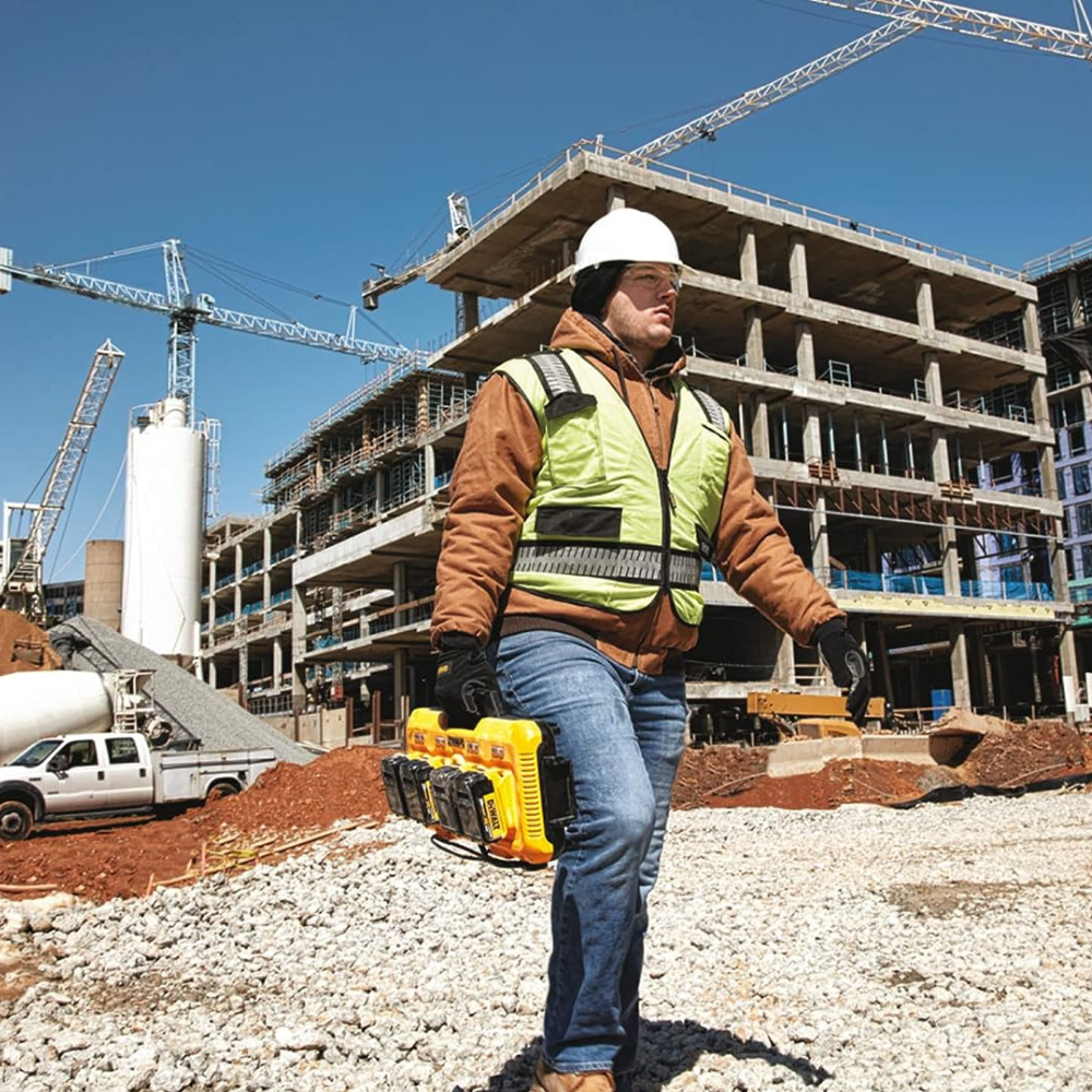 A worker carrying a 4 battery charger across a jobsite in gravel.