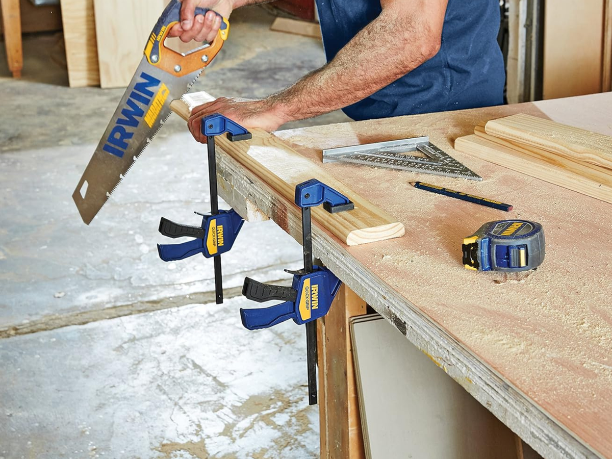 A man using an Irwin saw on a board held to a table with medium bar clamps.