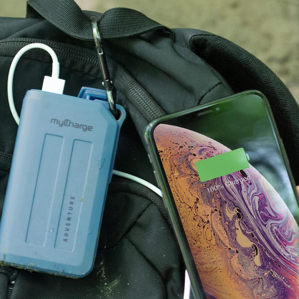 A rugged power bank hooked to a backpack and a phone showing 100% charge.