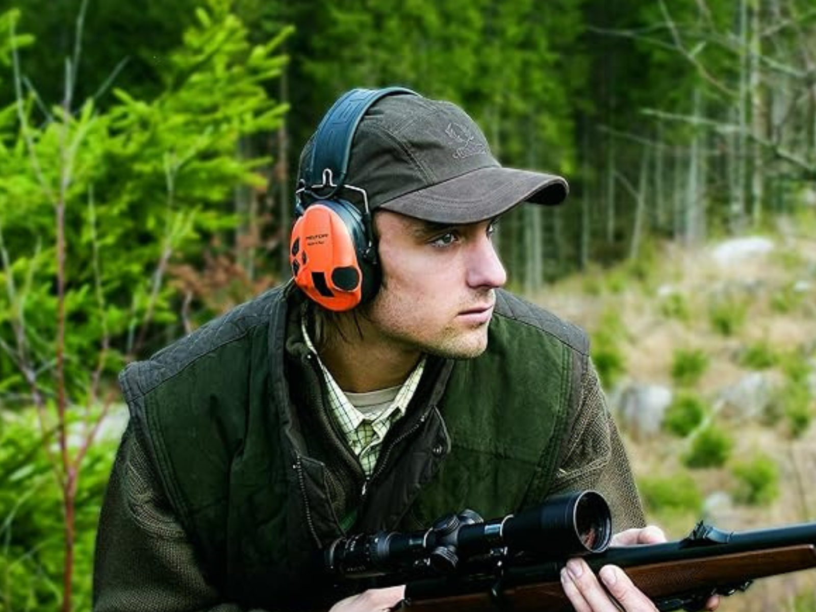 A man in hunter orange Peltor muffs with his rifle in the woods.